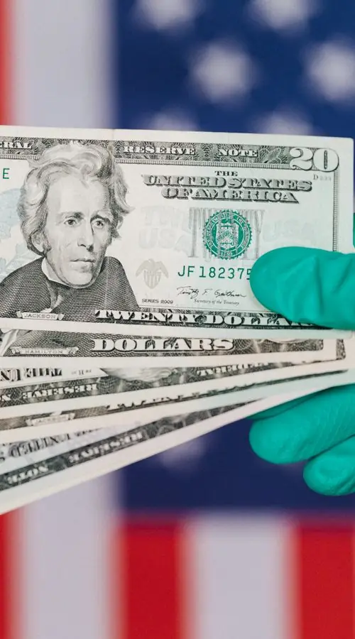 Gloved hand holding U.S. dollar bills in front of an American flag